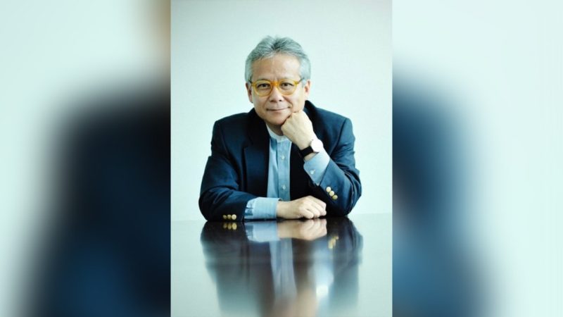 portrait of a middle-aged man with gray hair wearing yellow glasses, a blue shirt, and a dark blazer, sitting at a reflective table with his chin resting on his hand