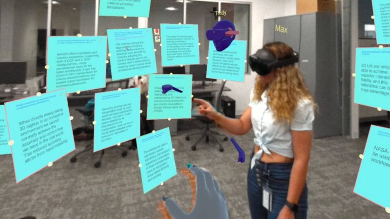 A person wearing a white shirt and bluejeans is seen from the side wearing an AR headset and clicking on one of 13 simulated notes floating around them. The notes are teal with black text which is not legible.